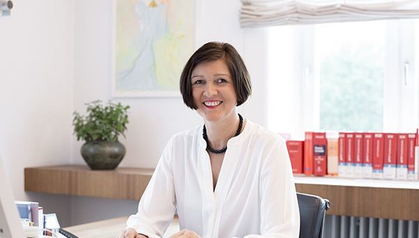 Eine Frau sitzt an einem Schreibtisch im Büro, lächelt und hält einen Aktenordner in den Händen.