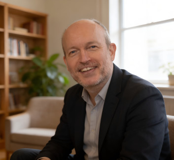 Lächelnder Mann in einem schwarzen Anzug in einem gemütlichen Raum mit Bücherregal und Pflanzen.