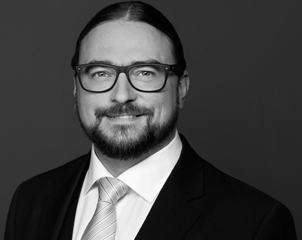 Porträt eines Mannes in formeller Kleidung mit Brille vor dunklem Hintergrund.