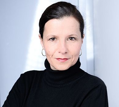 Porträt einer Frau mit dunklem Haar, die einen schwarzen Rollkragenpullover trägt, vor einem hellen Hintergrund.