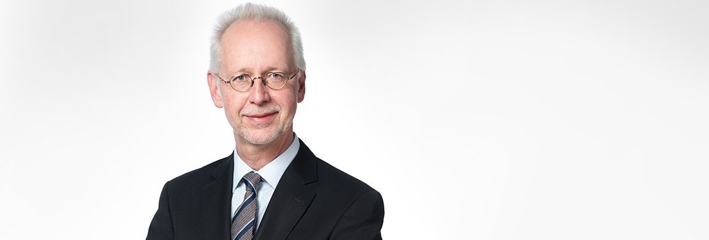 Ein älterer Mann in formeller Kleidung mit Anzug und Krawatte vor hellem Hintergrund.