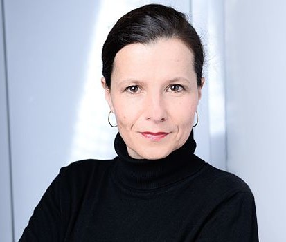 Porträt einer Frau mit dunklem Haar, die einen schwarzen Rollkragenpullover trägt, vor einem hellen Hintergrund.