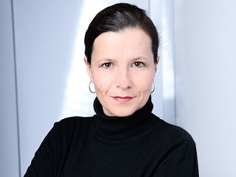 Porträt einer Frau mit dunklem Haar, die einen schwarzen Rollkragenpullover trägt, vor einem hellen Hintergrund.