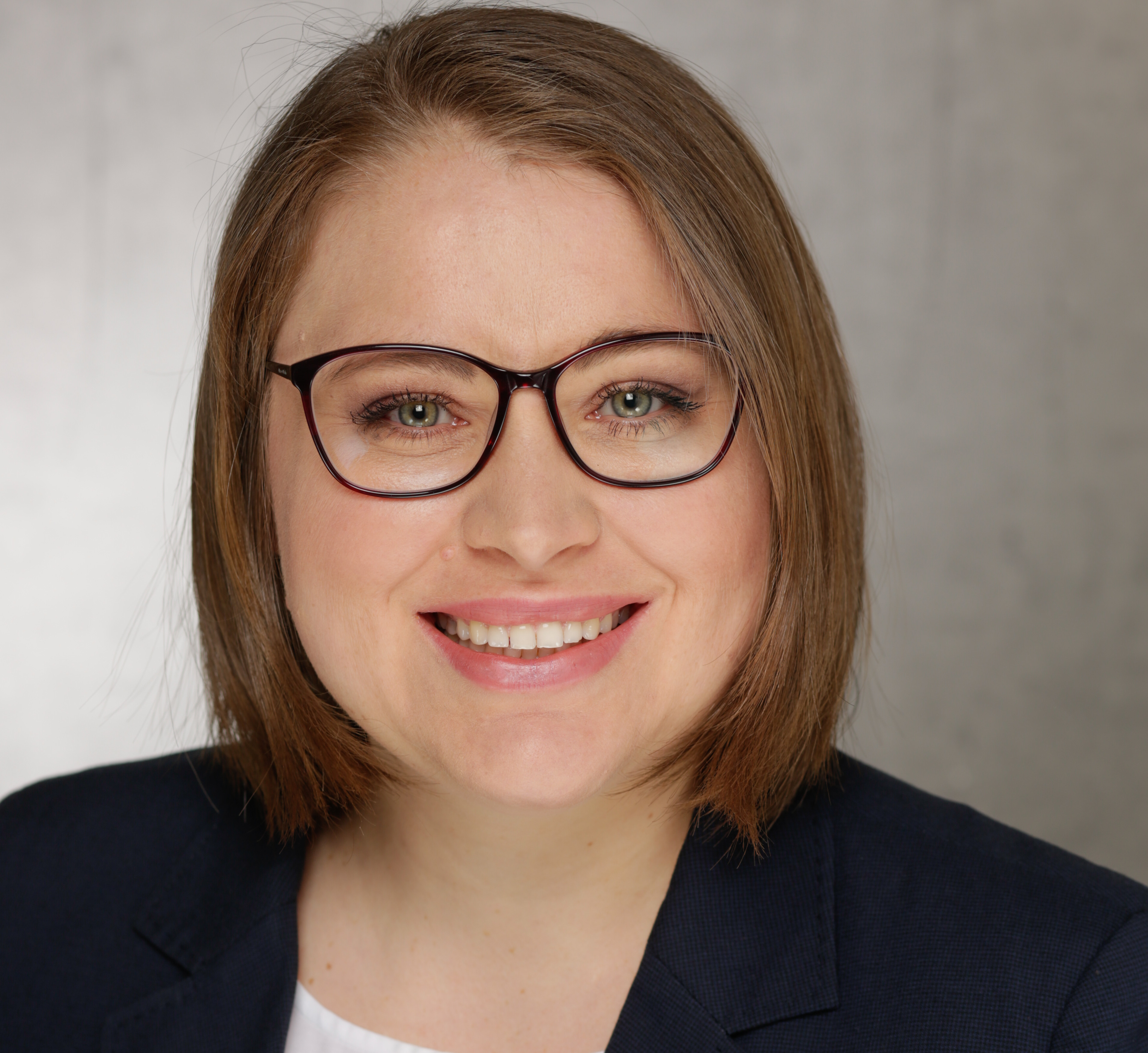 Porträt einer lächelnden Frau mit Brille in formeller Kleidung vor einem grauen Hintergrund.