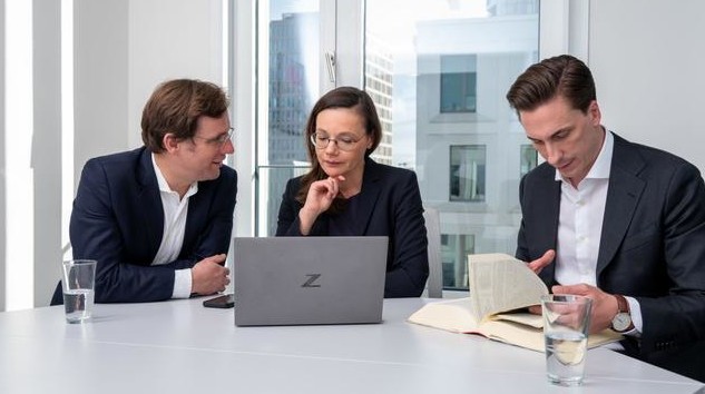 Drei Geschäftsleute bei einer Besprechung im Büro, einer liest in einem Buch, die anderen schauen auf einen Laptop.