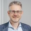 Lächelnder Mann in Brille, Business-Hemd und Blazer vor neutralem Hintergrund.