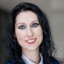 Porträt einer Frau mit schwarzen, lockigen Haaren und blauen Augen, lächelnd, im Business-Outfit.
