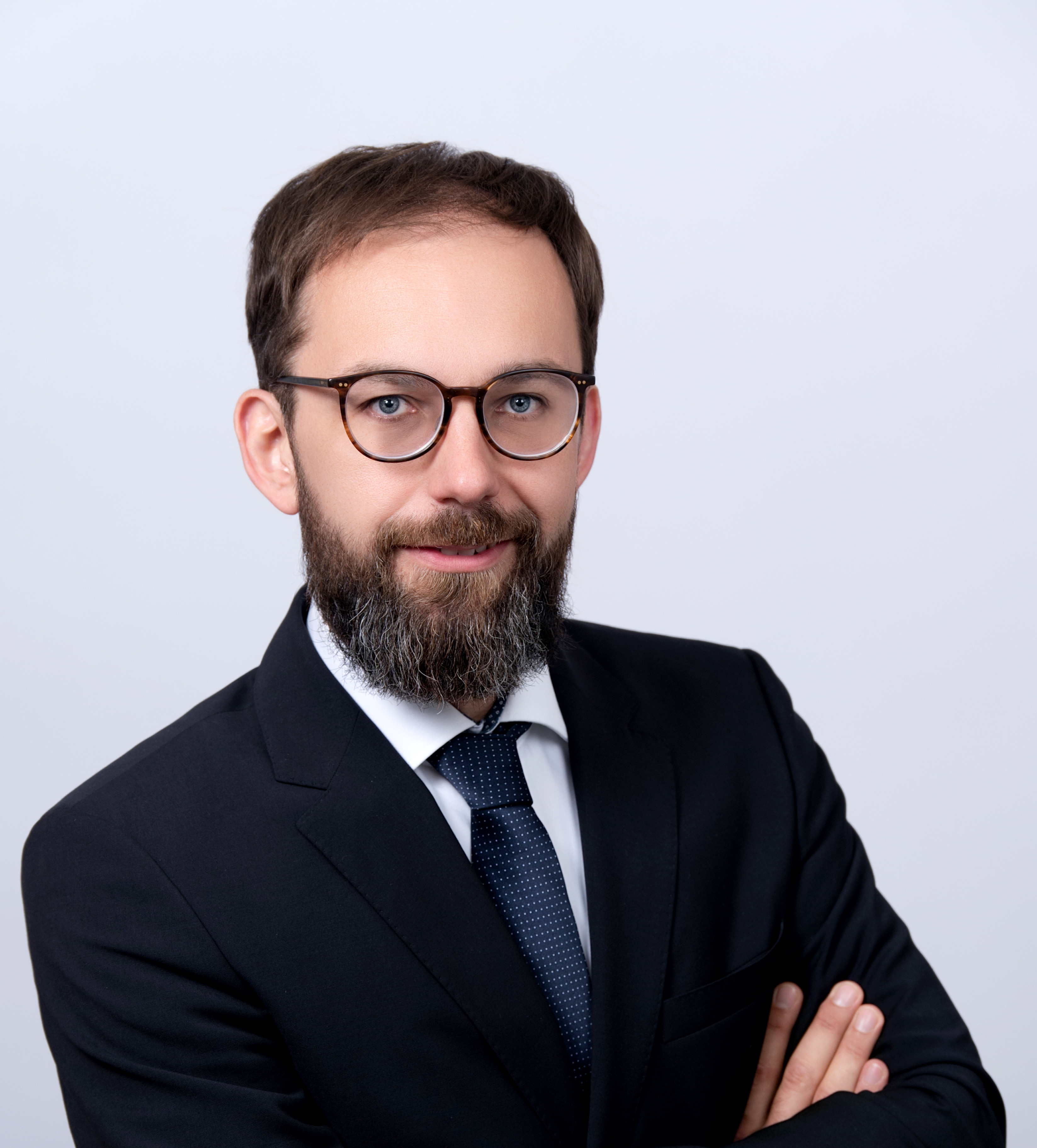 Porträt eines Mannes in formeller Kleidung mit Brille, Armbanduhr und gepflegtem Bart vor hellem Hintergrund.