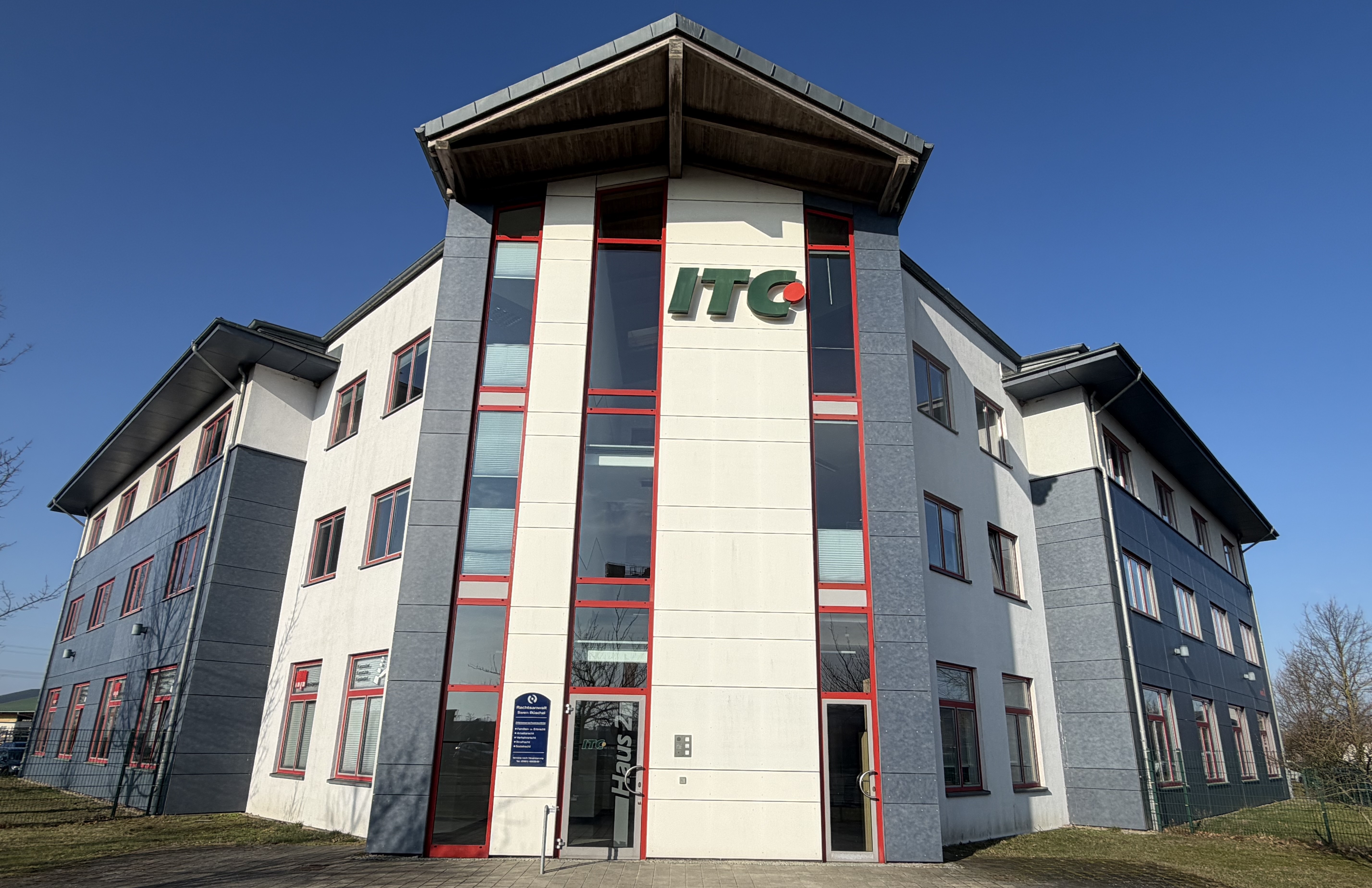 Mehrstöckiges Bürogebäude mit weißen und grauen Fassaden, rote Fensterrahmen und ein grünes und rotes "ITC" Logo an der Front.