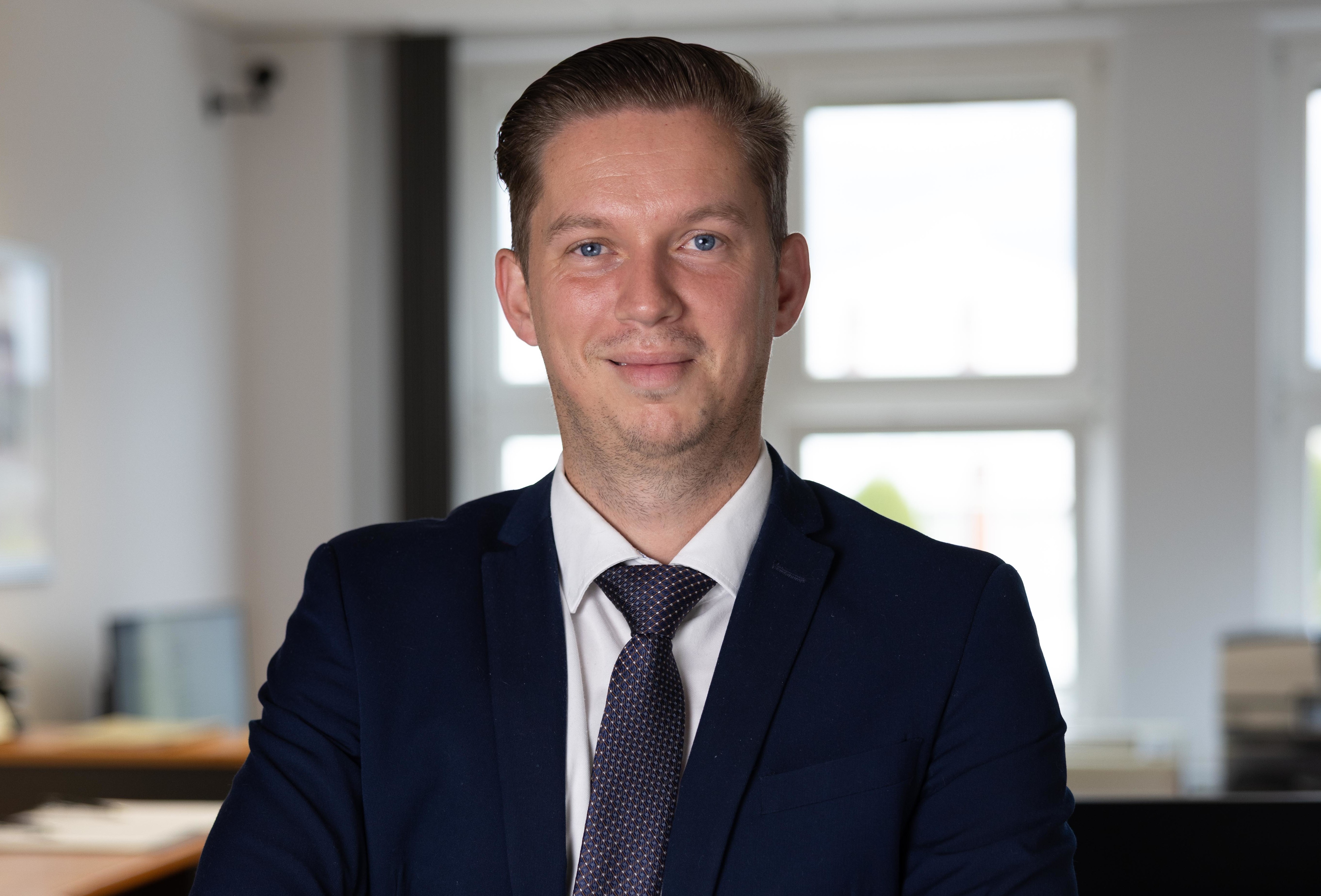 Ein Mann in einem dunklen Anzug mit Krawatte steht mit verschränkten Armen in einem Büro.