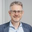 Lächelnder Mann in Brille, Business-Hemd und Blazer vor neutralem Hintergrund.