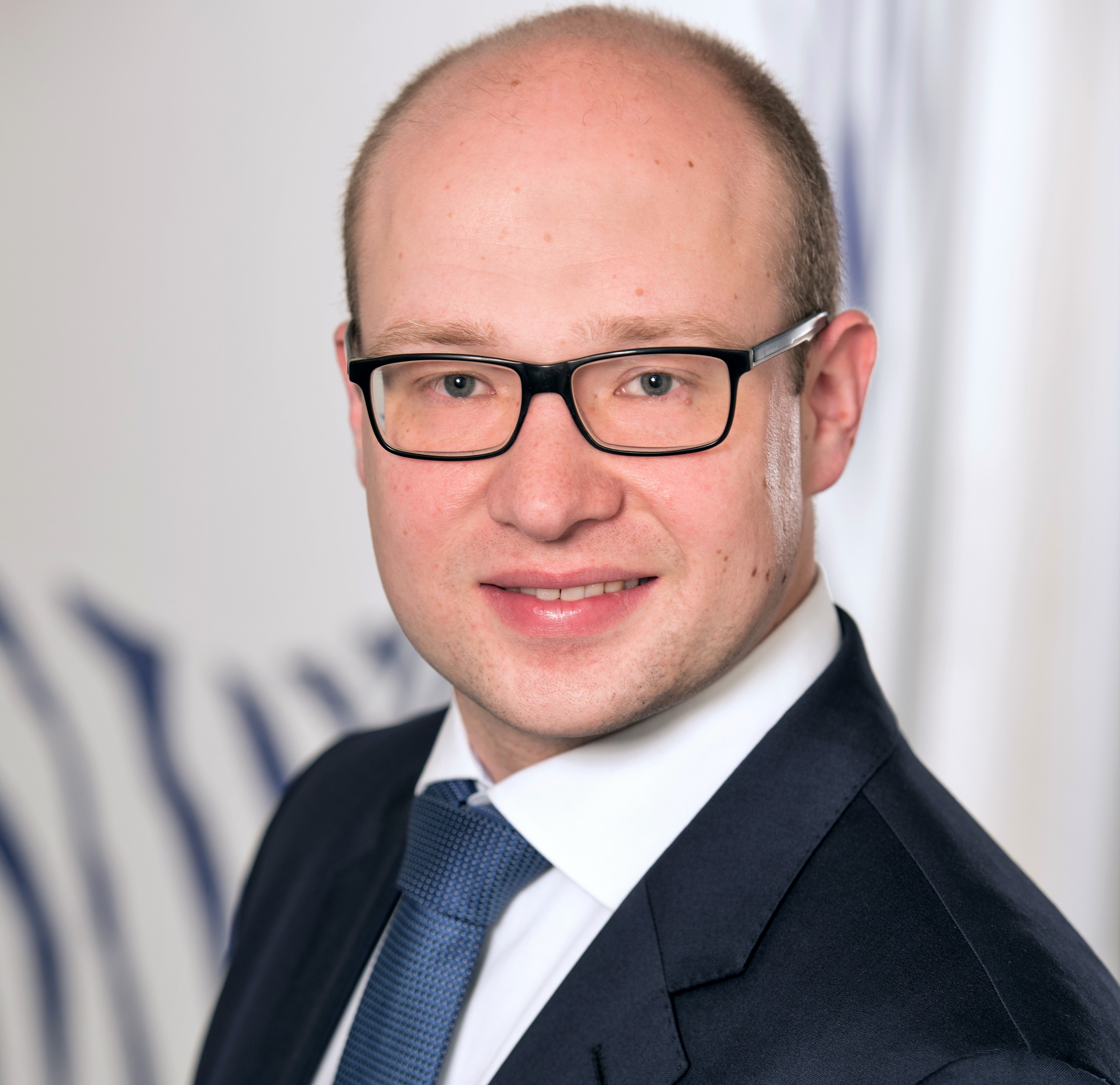 Porträt eines Mannes in formeller Kleidung mit Brille, lächelnd.