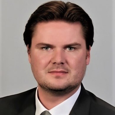 Porträt eines Mannes in formeller Kleidung, dunkler Anzug, weißes Hemd und graue Krawatte, vor hellem Hintergrund.