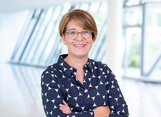 Eine lächelnde Frau mit Brille und kurzen Haaren steht in einem modernen, hellen Gebäude mit großen Fenstern.