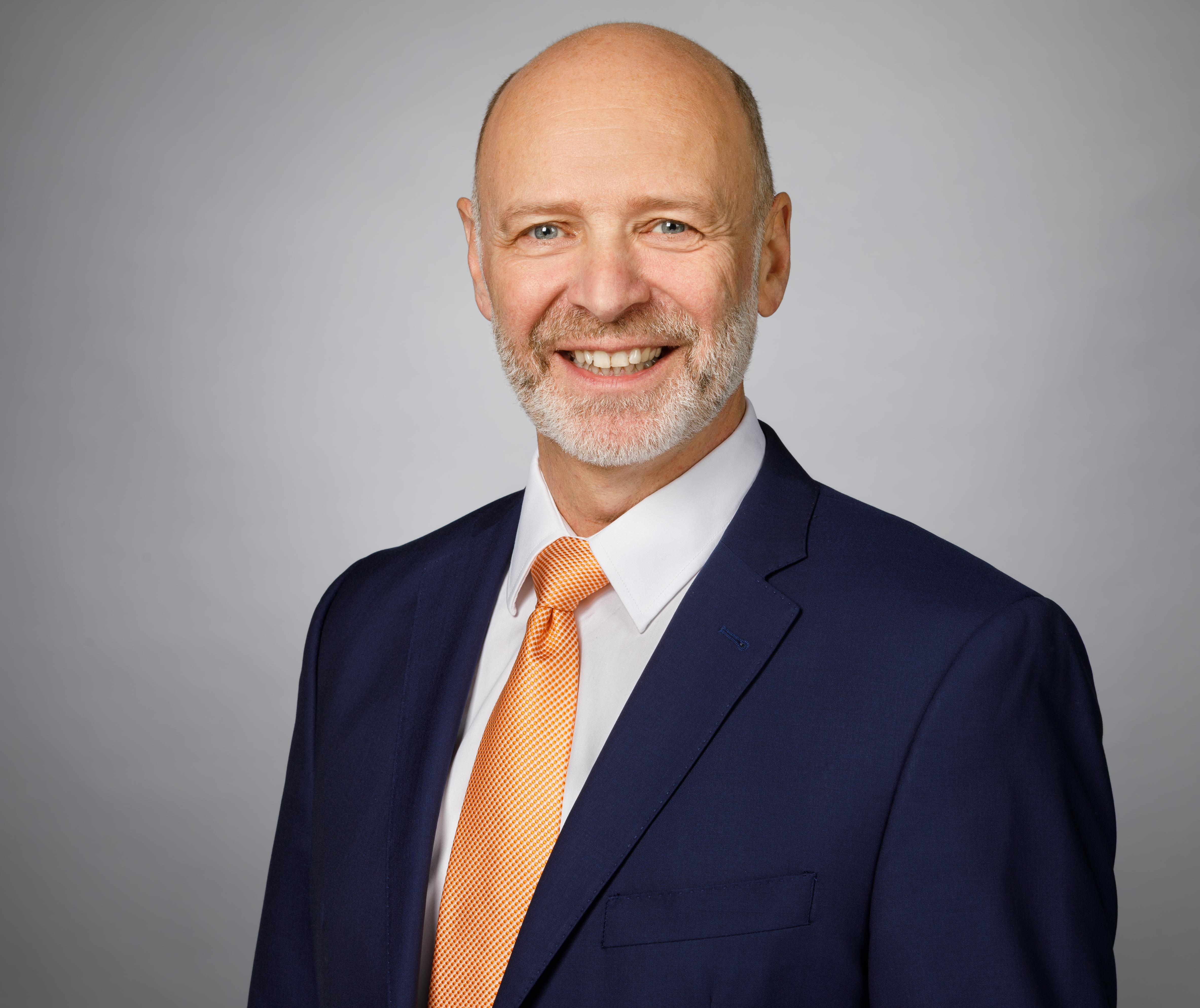 Mann in dunklem Anzug mit orangefarbenem Krawatte vor grauem Hintergrund.