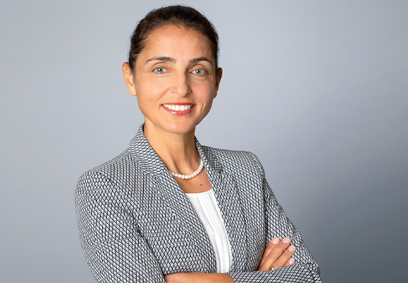 Eine professionell gekleidete Frau mit verschränkten Armen vor einem grauen Hintergrund.