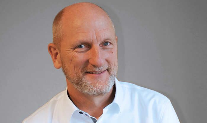 Ein lächelnder Mann mit kurzem, grauem Bart und blauen Augen, trägt ein weißes Hemd und steht vor einem grauen Hintergrund.