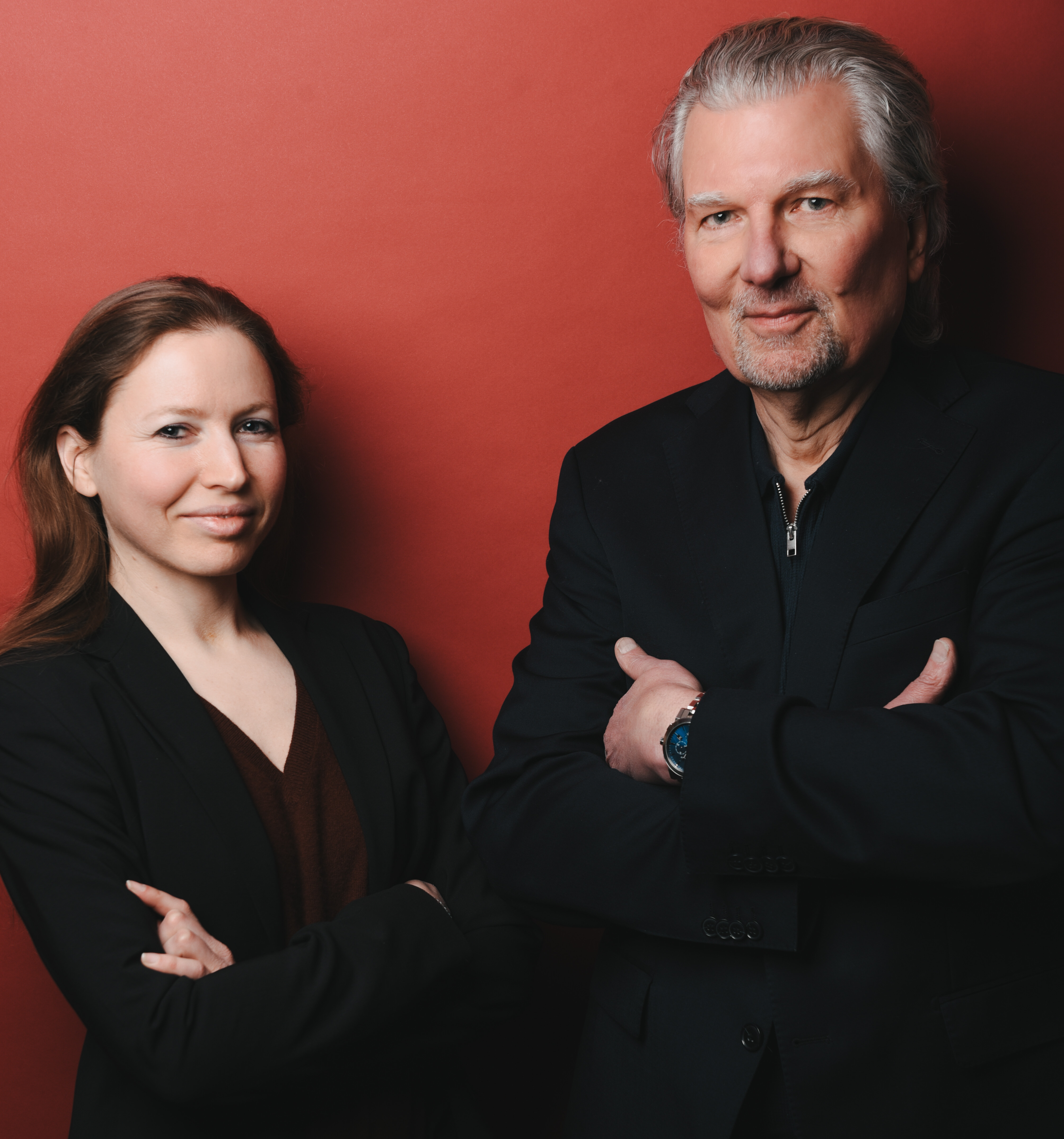 Zwei selbstbewusste Personen, eine Frau und ein Mann, stehen mit verschränkten Armen vor einem roten Hintergrund.