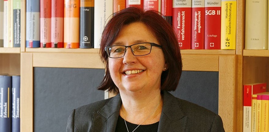 Frau mit Brille und dunklem Blazer vor einem Bücherregal mit bunten Büchern.