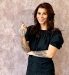 Friseurin mit Locken, Tattoos und Schere in der Hand vor einer rosa Wand, Logo "Iras Boudoir" im oberen linken Eck.
