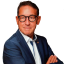 Porträt eines Mannes in einem blauen Anzug mit verschränkten Armen, trägt Brille und eine Armbanduhr.