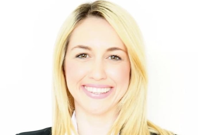 Porträt einer lächelnden Frau mit blonden Haaren, die formell gekleidet ist, vor weißem Hintergrund.