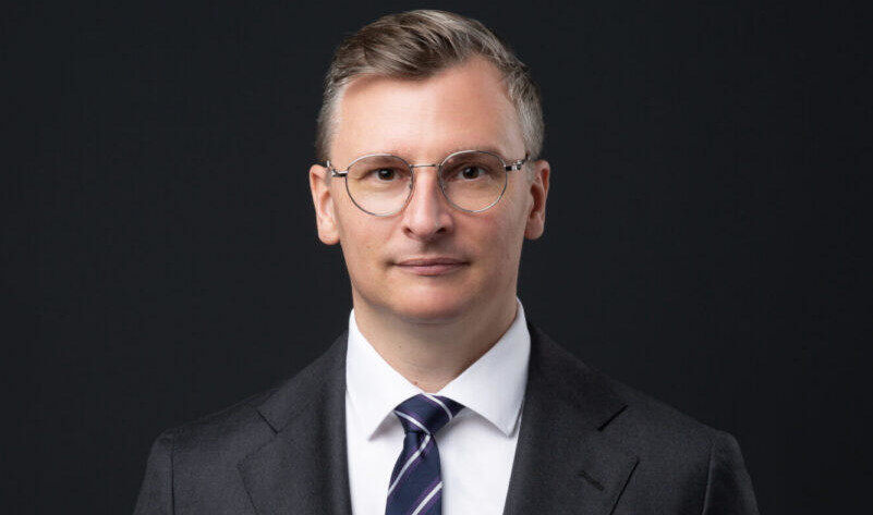Porträt eines Geschäftsmanns im dunklen Anzug mit gestreifter Krawatte und Brille vor schwarzem Hintergrund.