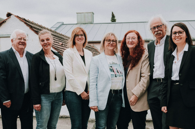 Gruppe von acht Menschen, die draußen vor einem Gebäude stehen und lächeln.