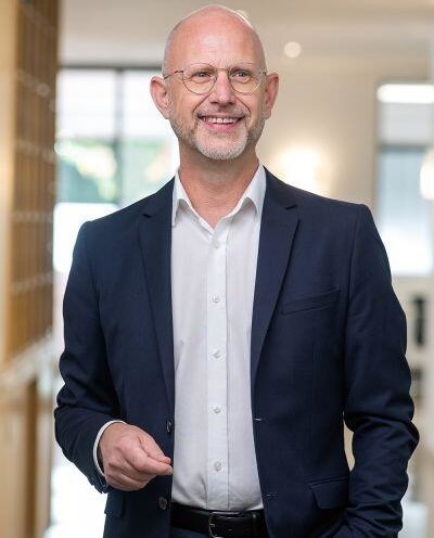 Lächelnder Mann in formeller Kleidung vor hellem, modernem Hintergrund.