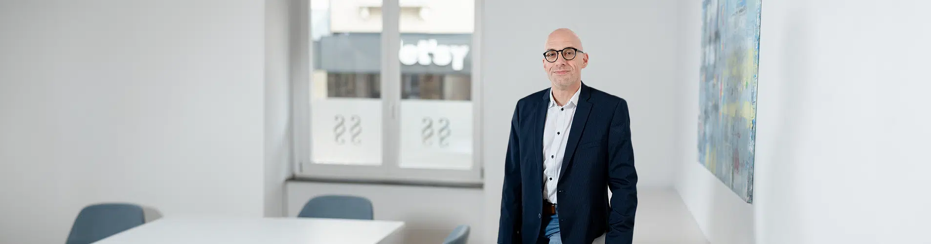 Ein Mann in einem dunklen Anzug steht in einem modernen, hellen Büro vor einer weißen Wand.