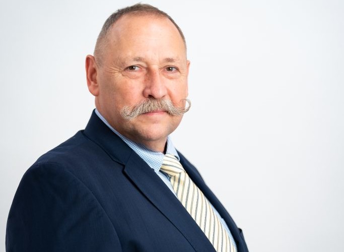 Ein Mann mit Schnurrbart in einem dunklen Anzug und Krawatte posiert vor weißem Hintergrund.