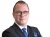 Ein Mann in einem schwarzen Anzug mit Brille, blauer Krawatte und Armbanduhr, der die Arme verschränkt.
