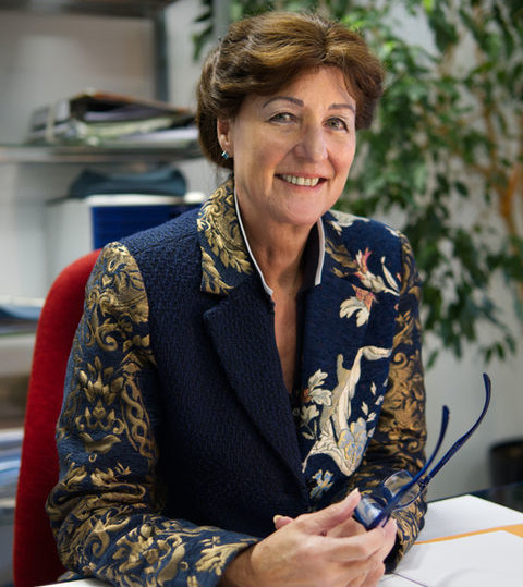 Frau sitzt an einem Schreibtisch im Büro, lächelt und hält eine Brille in der Hand.