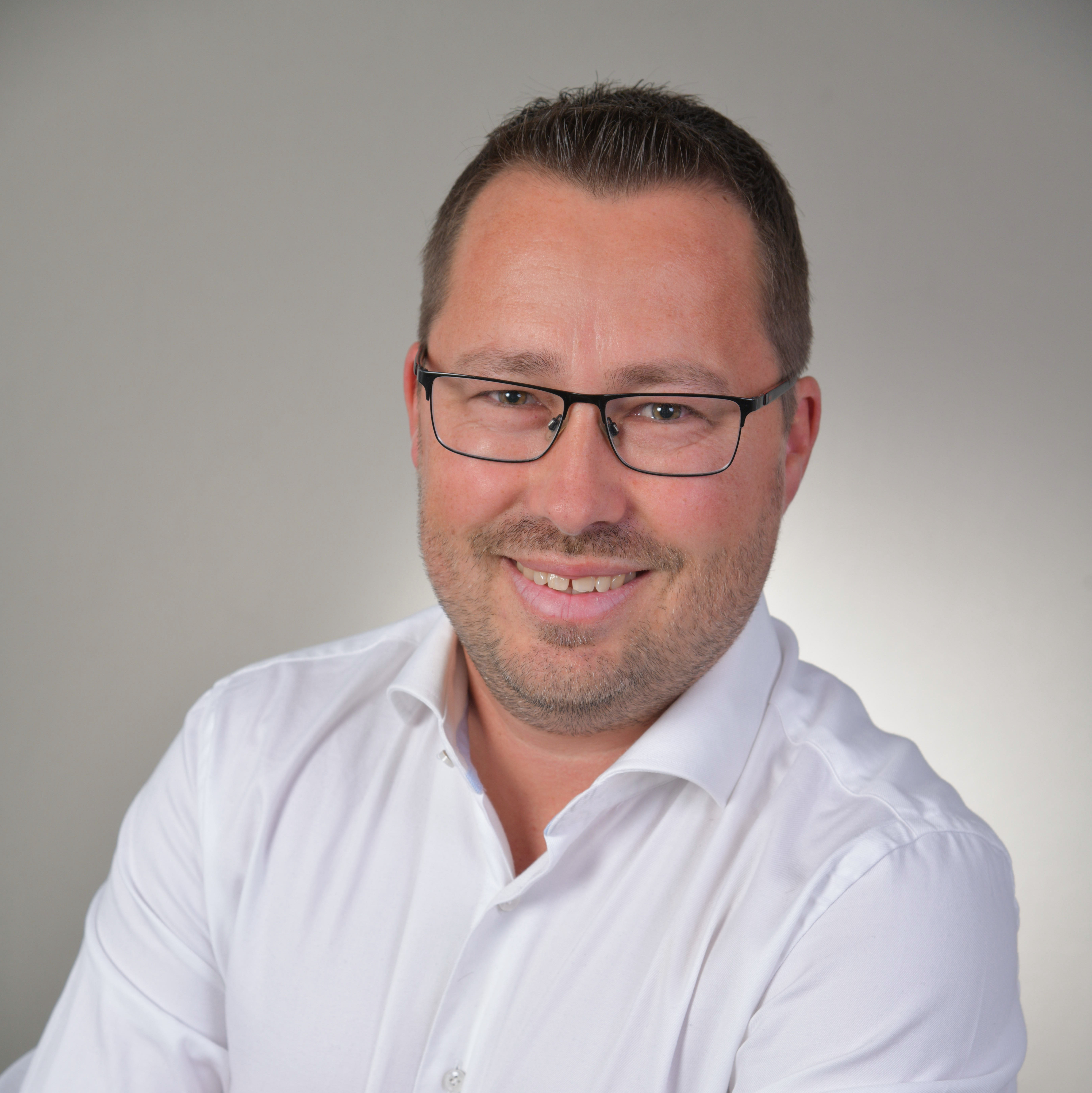 Lächelnder Mann mit Brille in weißem Hemd, Arme verschränkt, vor neutralem Hintergrund.