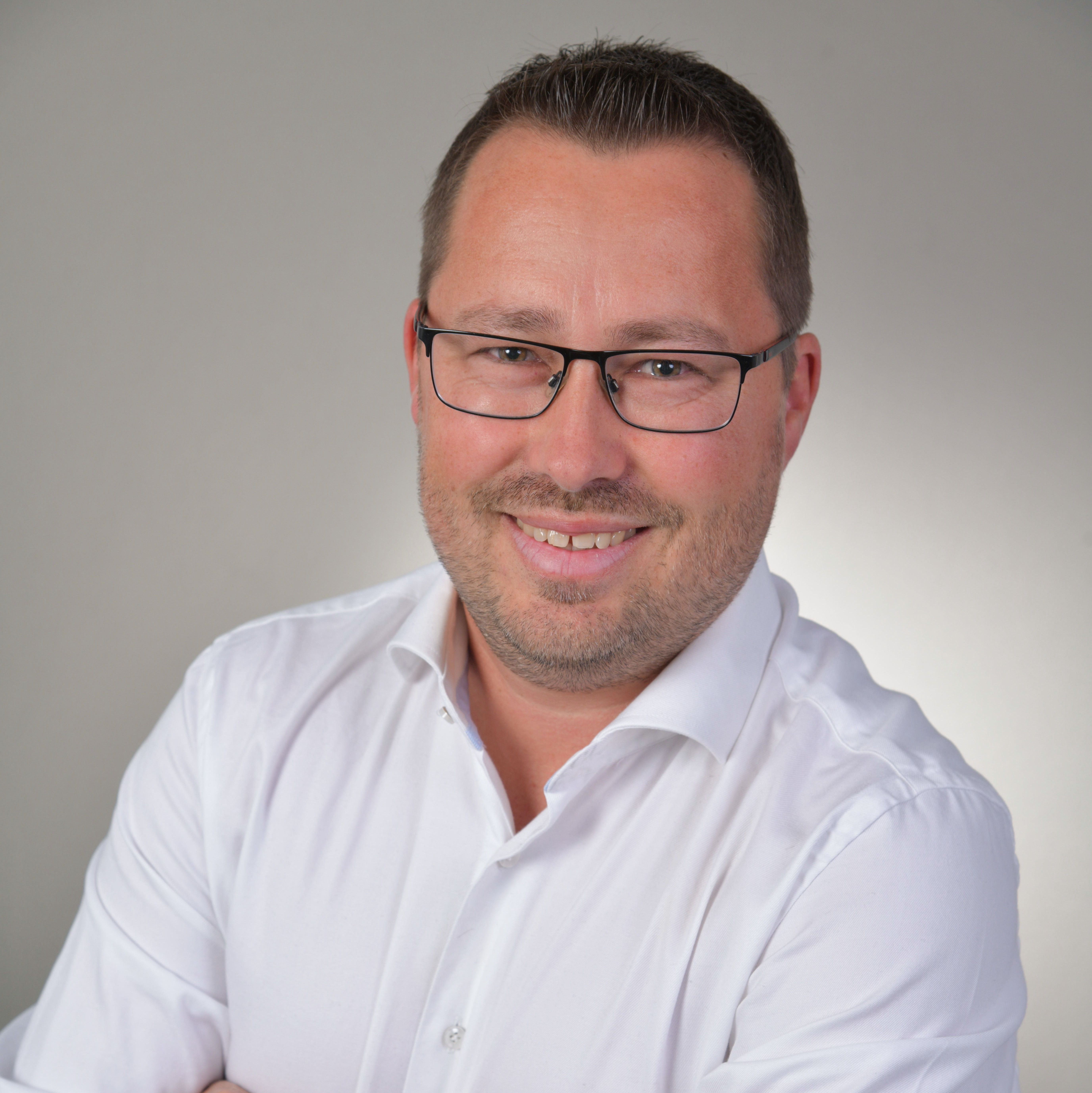 Lächelnder Mann mit Brille in weißem Hemd, Arme verschränkt, vor neutralem Hintergrund.