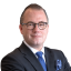 Ein Mann in einem schwarzen Anzug mit Brille, blauer Krawatte und Armbanduhr, der die Arme verschränkt.