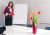 Frau steht vor Whiteboard und Flipchart, hält Notizen in der Hand, während sie eine Präsentation hält, im Konferenzraum mit Blumen im Vordergrund.