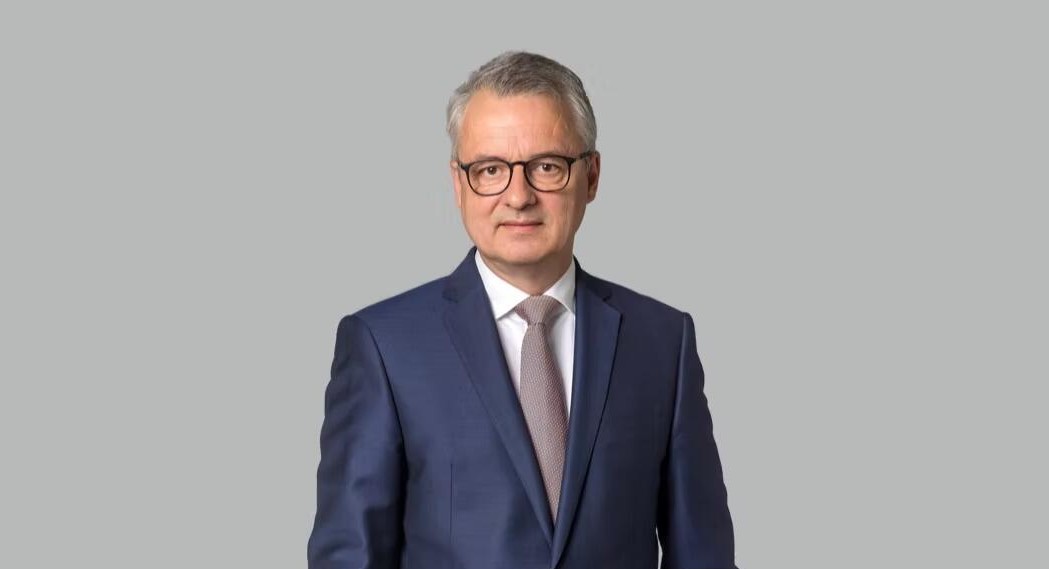 Ein Mann in einem dunklen Anzug mit Krawatte vor einem grauen Hintergrund.