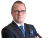 Ein Mann in einem schwarzen Anzug mit Brille, blauer Krawatte und Armbanduhr, der die Arme verschränkt.