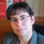 Porträt einer Frau mit dunklem Haar, Brille mit rosa Rahmen, grauem Blazer, vor einem Bücherregal.