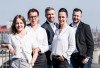 Eine Gruppe von fünf professionell gekleideten Menschen steht auf einer Brücke mit Stadtblick im Hintergrund.