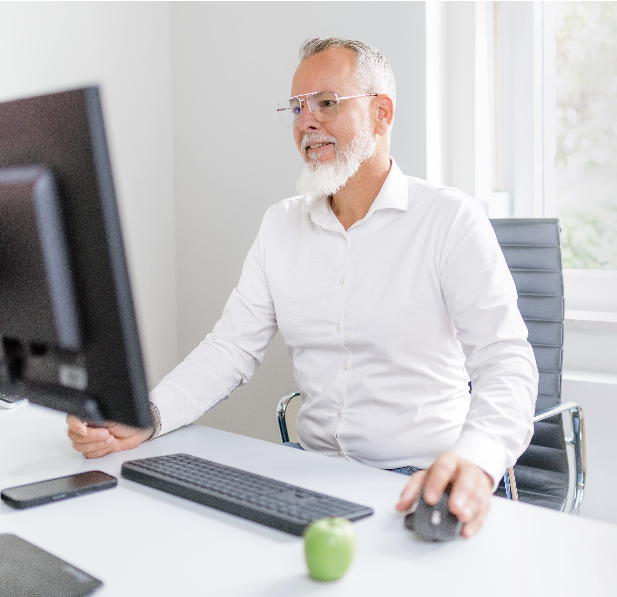 Ein älterer Mann mit Brille und grauem Bart arbeitet an einem Schreibtisch im Büro, vor einem Computer.