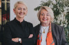 Zwei lachende Frauen stehen nebeneinander vor einer Pflanze und einem weißen Hintergrund.