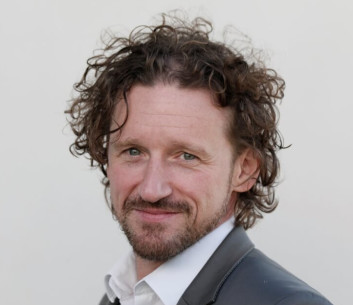 Porträt eines Mannes mit lockigem Haar und Bart, der einen schwarzen Anzug trägt.