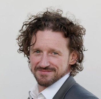 Porträt eines Mannes mit lockigem Haar und Bart, der einen schwarzen Anzug trägt.