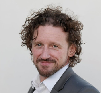 Porträt eines Mannes mit lockigem Haar und Bart, der einen schwarzen Anzug trägt.