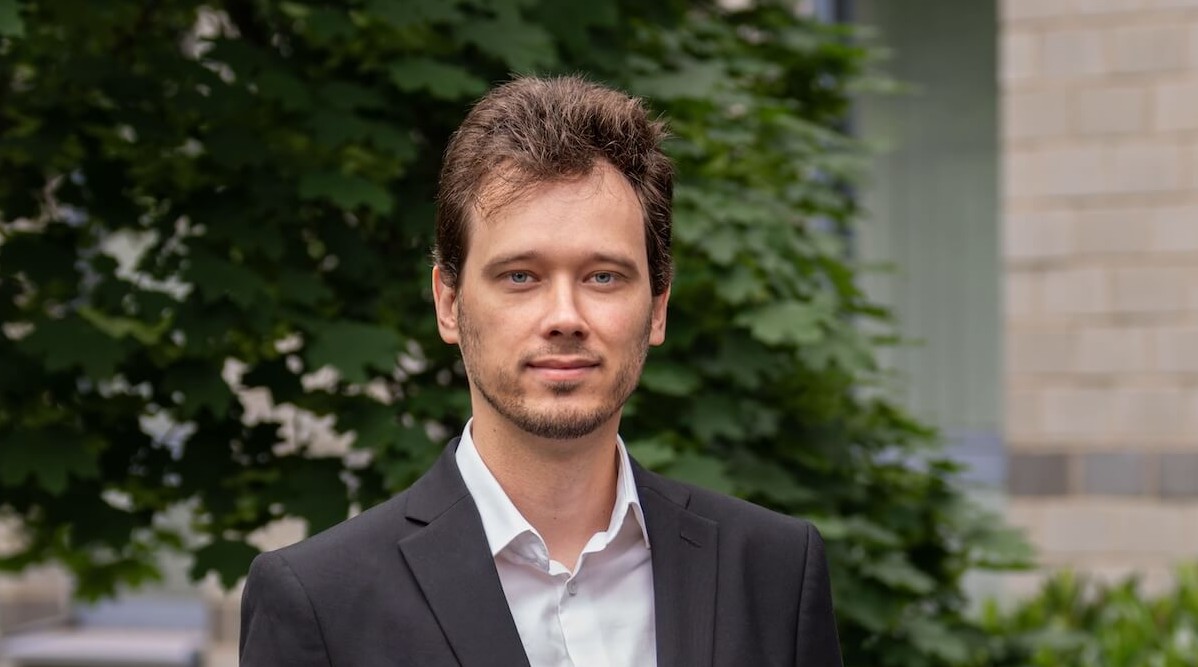 Junger Mann in formellem schwarzen Anzug vor grüner Hecke und Gebäudewand.