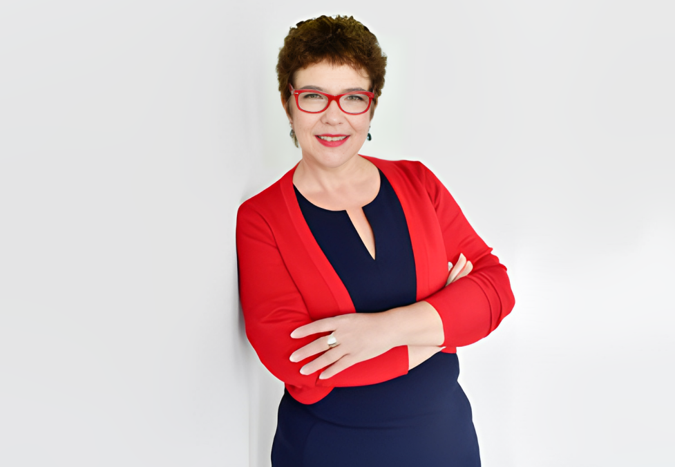 Frau mit roten Brillen und rotem Blazer, die lächelnd mit verschränkten Armen vor weißem Hintergrund steht.