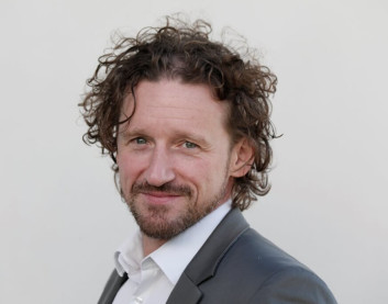 Porträt eines Mannes mit lockigem Haar und Bart, der einen schwarzen Anzug trägt.