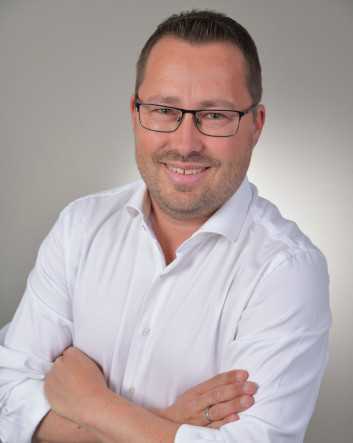 Lächelnder Mann mit Brille in weißem Hemd, Arme verschränkt, vor neutralem Hintergrund.